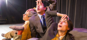 A photo of three cast members of Santa Fe's Theater Grottesco during a scene of the play The Moment of YES! In the photo, two men and a woman make dramatic faces while sprawled on a stage floor.