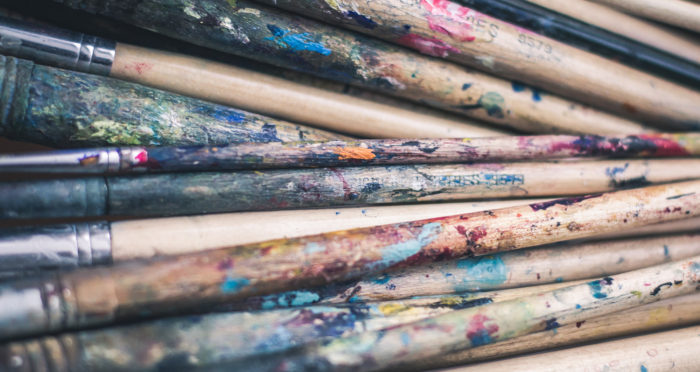 A closeup photograph of the wood handles of dozens of paintbrushes. The handles are covered in various dried paint colors.
