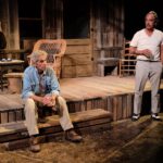 A picture of two men performing a scene from Ages Of The Moon By Sam Shepard at Teatro Paraguas Theater, Santa Fe, New Mexico. The men are in front of a wood house. The man on the left is sitting on the porch steps, while the man on the right, holding a glass, is looking at him.