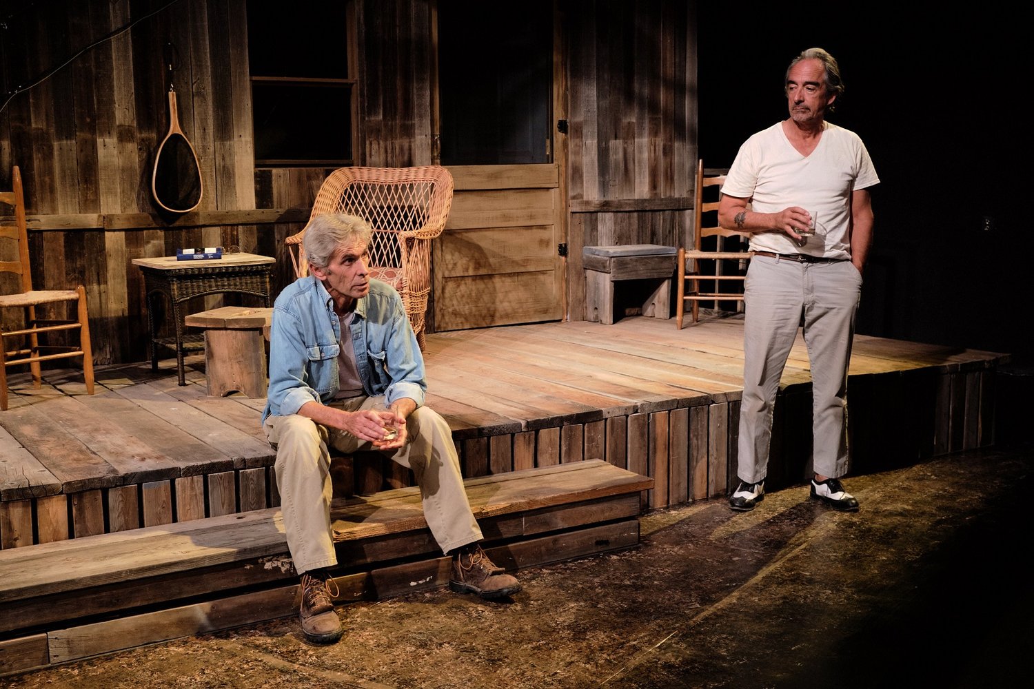 A picture of two men performing a scene from Ages Of The Moon By Sam Shepard at Teatro Paraguas Theater, Santa Fe, New Mexico. The men are in front of a wood house. The man on the left is sitting on the porch steps, while the man on the right, holding a glass, is looking at him.