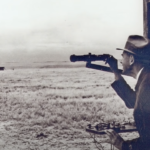 An old, black and white image of a man using a scope to look out over a field in Roswell, NM.