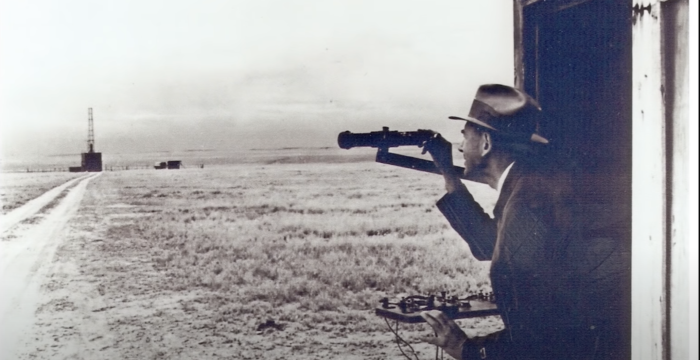 An old, black and white image of a man using a scope to look out over a field in Roswell, NM.