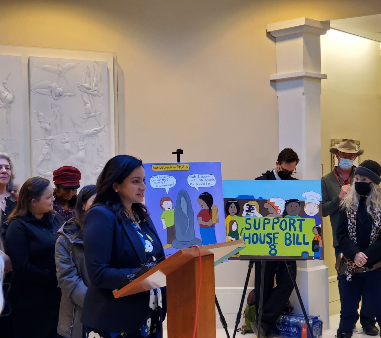 A New Mexico state legislator stands at a podium in the atrium of the state capitol. Around her are supporters of NM House Bill 8, which would support the arts in New Mexico.