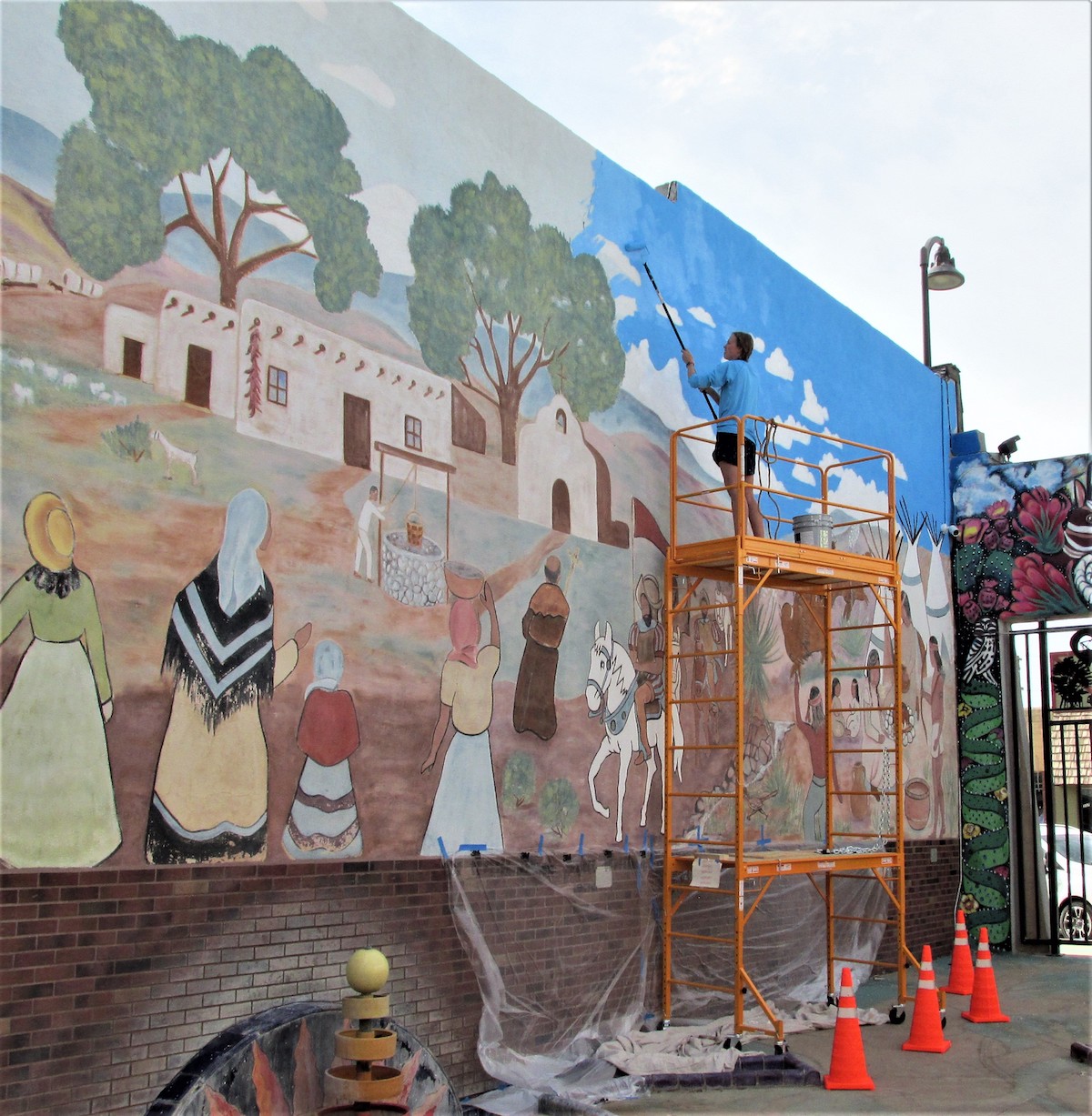 A woman stands on a lift, painting blue onto the sky portion of a mural. The mural, in Artesia NM, depicts the square of a small adobe village from colonial times.
